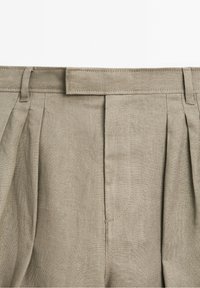 Ceinture de pantalon en lin beige montrant des passants de ceinture, des plis et une fermeture à crochet et barre recouverte de tissu.