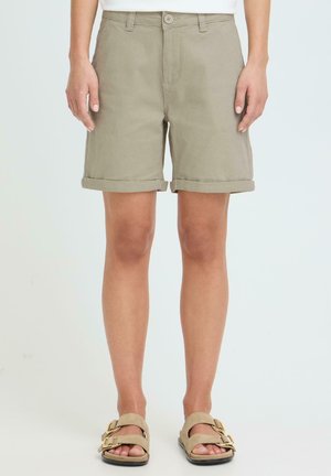 Person kledd i beige shorts med oppbrettede kanter, stående med armene avslappet, kombinert med beige sandaler med spenne mot en ensfarget bakgrunn.