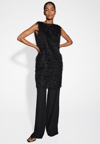 Abito nero senza maniche con un design texturizzato e frangiato, abbinato a pantaloni neri larghi e sandali bassi, su uno sfondo bianco.