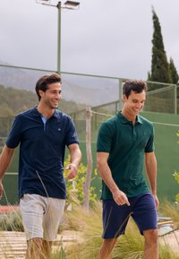 Två män går utomhus klädda i kortärmade polo-tröjor; en i marinblått med en ljusblå krage och den andra i mörkgrön, tillsammans med shorts.