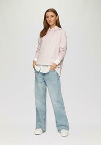 Felpa con cappuccio rosa chiaro, maniche lunghe, indossata sopra una camicia bianca. Abbinata a jeans azzurri a gamba larga e sneakers bianche.