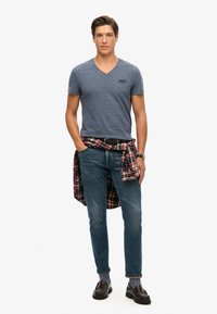 T-shirt blu con scollo a V e piccolo logo, jeans scuri e camicia a quadri annodata in vita; mocassini marroni completano l'outfit.