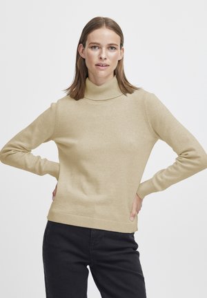 Pull over beige clair en maille douce avec col roulé, présentant des poignets et un ourlet à côtes. Porté avec un pantalon noir ; le tissu semble lisse et chaud.
