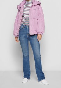 Hellviolette Steppjacke mit Kapuze, gesteppter Schnitt; getragen über einem gestreiften Langarmshirt, kombiniert mit ausgestellten blauen Jeans und weißen Turnschuhen.