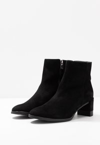 Bottines en suède noir avec un bout pointu, une fermeture éclair sur le côté et un talon carré. La texture est douce et le design est élégant et minimaliste.