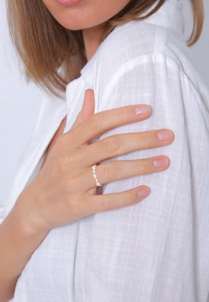 Main de femme avec une bague en perle reposant sur l'épaule d'une chemise en tissu blanc texturé.
