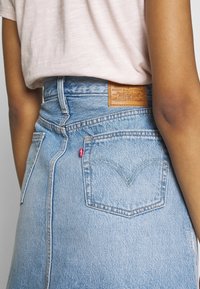 Světle modrá džínová sukně s vysokým pasem, obsahující dvě zadní kapsy a koženou značkovou etiketu na pásku. Prošité detaily v žluté barvě.