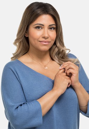 Blauer, gestrickter Pullover mit V-Ausschnitt und Dreiviertelärmeln. Die Anhänger-Kette hat ein strukturiertes Blatten-Detail, die Hautfarbe ist an den Händen sichtbar.