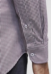 Nahaufnahme des Arms einer Person, die ein langärmliges, lila-weiß kariertes Hemd mit geknöpften Manschetten und schwarze Hosen trägt.