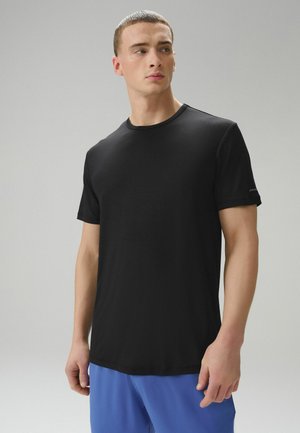 Ung mand med kort hår iført en sort t-shirt med korte ærmer og blå bukser, der kigger lidt til sin venstre side på en ens grå baggrund.