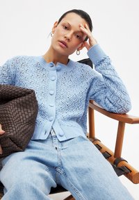 Svetlo modri pleteni kardigan z čipkastim vzorcem, s krožnimi gumbi in rebrastimi manšetami, v kombinaciji s svetlimi kavbojkami in rjavim tkanim žakom.