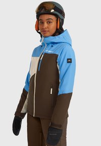 Zila, brūna un balta slēpošanas jaka ar rāvējslēdzēja aizdari, regulējamu kapuci un sānu kabatām. Melnas cimdi ar logotipa detaļu, attēloti uz neitrāla fona.