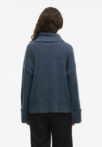 Pull en maille texturée bleu foncé, surdimensionné, avec un col haut et replié et des manches larges. Base courte avec des bords bruts.
