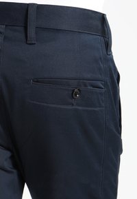 Tmavě modré kalhoty s hladkou texturou, s zadní kapsou zabezpečenou černým knoflíkem a střihem s přímým střihem.