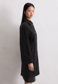 Cappotto in lana nera con colletto, maniche lunghe, due tasche anteriori e chiusura con bottoni, indossato da un modello su uno sfondo neutro.
