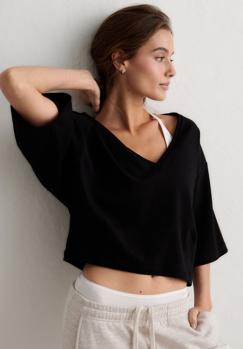 Tânără îmbrăcată într-un top negru larg, bustieră sport albă și pantaloni de trening deschiși la culoare, pozând pe un perete alb, uitându-se spre dreapta cu mâna pusă în spatele capului.