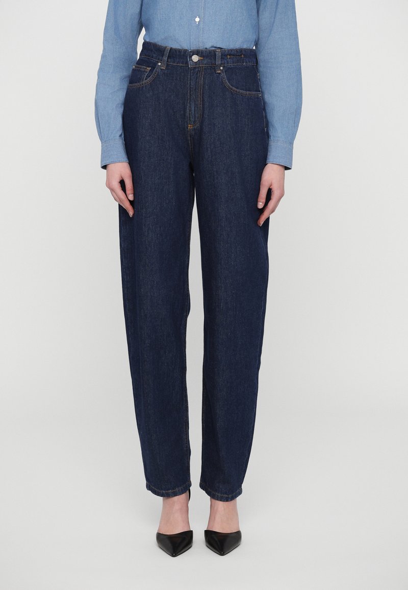 Mørkeblå denimjeans med høy midje, rette ben, kontrasterende oransje sømmer og frontlommer. Kombinert med svarte spisse sko.