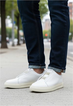 Persona con jeans azul oscuro remangados y zapatillas blancas sin cordones, de pie en una acera de la ciudad.