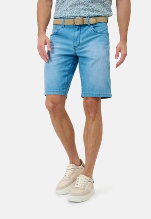 Homme portant un short en jean bleu clair, une ceinture beige, une chemise à motifs et des baskets beiges, debout sur un fond blanc.