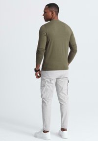 Mann trägt ein olivgrünes Langarmshirt, hellgraue Cargo-Hose, weiße Sneakers und eine schwarze Armbanduhr, steht seitlich gerichtet.
