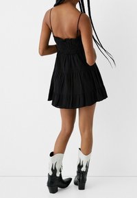 Svart, lager-på-lager mini-klänning med tunna axelband och rynkad livstycke. Kombinerad med svarta cowboystövlar med vita flammande detaljer.