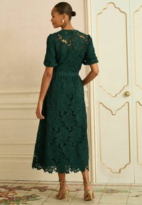 Zaļš mežģīņu kleita ar ziedu rakstiem, elkoņa garuma piedurknēm un vidējas garuma apakšmalu. Aizmugurē aizdares ar rāvējslēdzēju un kombinēta ar papēžu kurpēm.