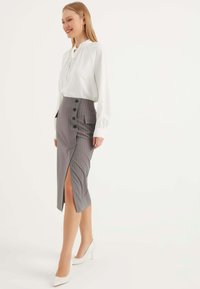 Weiße Bluse mit Rüschenausschnitt, kombiniert mit einem grauen Bleistiftrock mit Nadelstreifen, der einen seitlichen Schlitz und schwarze Knopfdetails aufweist. Weiße Pumps.