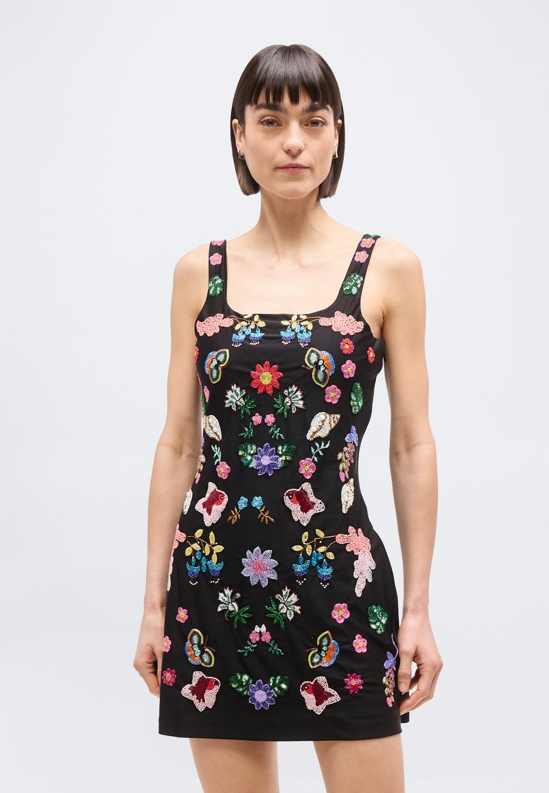 Kvinde iført en sort ærmeløs kjole med farverige broderede blomster og sommerfugle, stående mod en ensfarvet lys baggrund.