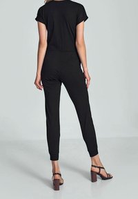 Combinaison noire à manches courtes, taille élastique, jambes fuselées et texture lisse. Associée à des sandales à talons blocs marron.