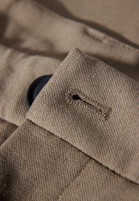 Beżowa tkanina posiada dziurkę na guzik oraz czarny guzik, z widocznym przeszyciem i teksturowaną powierzchnią. Projekt podkreśla dopracowane detale.