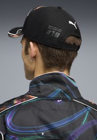 Schwarze Kappe mit buntem abstraktem Design, ausgestattet mit einem gebogenen Schirm, einem verstellbaren Riemen und Logodetail, kombiniert mit einer gemusterten Jacke.