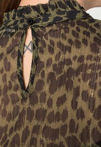 Blouse en tissu transparent à imprimé animal marron et vert avec un col volanté. Présente un bouton noir à l'arrière avec une ouverture en trou de serrure.