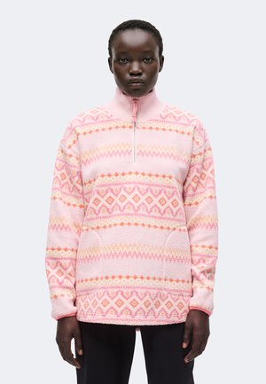 Personne à la peau foncée portant un pull polaire rose clair avec des motifs géométriques orange et roses et une fermeture éclair à l'avant, debout devant un fond uni.
