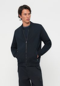 Veste bomber zippée bleu marine en coton doux. Dispose d'un col montant, de longues manches et de poches latérales à l'avant pour plus de praticité.