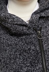 Veste à capuche en tricot gris avec une surface texturée. Comprend une fermeture éclair noire et un ajustement près du cou, conçue pour la chaleur et le confort.