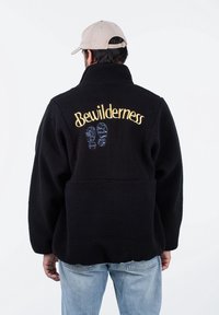 Czarna kurtka polarowa z wysokim kołnierzem, z haftem napisu "Bewilderness" w kolorze złoto-niebieskim oraz niebieskimi odciskami butów na plecach.