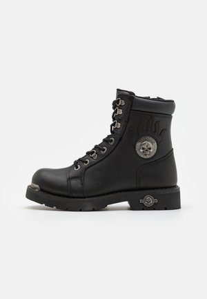 Botas de cuero negro hasta el tobillo con un cuello acolchado, ojales de metal, un logo de calavera y una suela de goma resistente. Cuenta con puntera y talón reforzados.