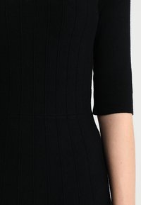 Robe en maille noire avec une texture côtelée, taille ajustée et manches courtes. Le tissu est lisse avec une légère élasticité. Aucune quincaillerie visible.