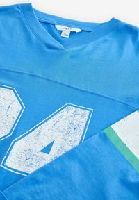 Sininen puuvillaneule, jossa on V-kaula-aukko, suuri valkoinen kulunut numero "94" edessä ja vihreä korosteraidassa hihassa.