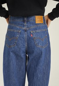 Blå denimjeans med rak bendesign, featuring två bakfickor, en läderlapp vid midjan och kontrasterande gula sömmar.