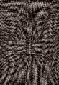 Brązowa tkanina w wzór houndstooth z fakturowanym splotem, z paskiem w talii i plisami poniżej, tworząca dopasowany wygląd.