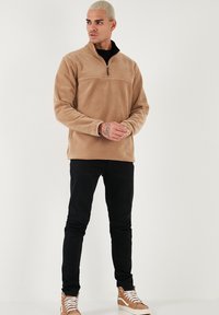 Pullover in pile color beige con colletto a mezza zip, maniche lunghe e una texture liscia, abbinato a pantaloni neri e sneakers beige.