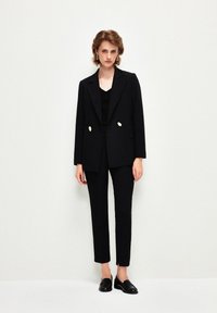 Černý šitý blazer s dvojitými knoflíky, zkombinovaný s úzkými černými kalhotami. Model má na sobě černý top a mokasíny, na bílém pozadí.