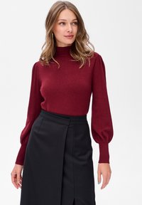 Kobieta z falującymi, jasnobrązowymi włosami nosi bordową bluzkę z długim rękawem i bufkami oraz czarną spódnicę o wysokim stanem z rozporkiem z przodu.