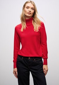 Maglione a collo alto rosso realizzato in tessuto a maglia, con maniche lunghe e design aderente. Abbinato a jeans di denim blu scuro.