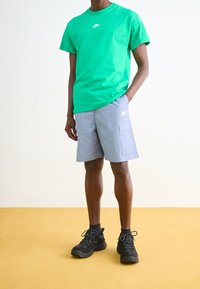 Grön bomullströja med vitt tryck, grå cargoshorts med sidofickor och svarta träningsskor. Enkel bakgrund med gul golv.