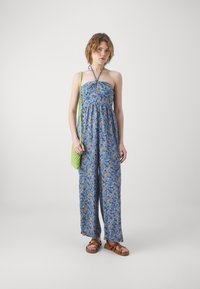 Modra cvetna kombinezona z vrvico okoli vratu, zbranim pasom, širokimi hlačami in zeleno tkanino torbo. Rjave sandale s trakovi dopolnjujejo videz.