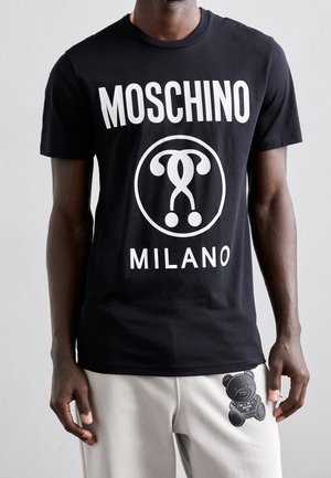 Man wearing black Moschino Milano T-shirt with white logo and light pants featuring black teddy bear graphic on thigh.