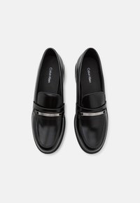 Mocassins en cuir noir avec une conception plate, une silhouette élégante et une bande d'accent métallique portant le logo de la marque sur le devant.