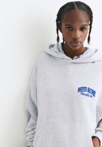 Grå hoodie lavet af blødt materiale, med en frontlomme og blåt broderet tekst "NEED MORE Vacation." Casual, løs pasform.